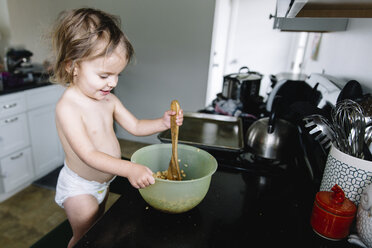 Junges Mädchen in Windel steht auf einem Hocker in einer Küche, hält einen Holzlöffel und rührt Essen in einer Plastikschüssel um. - MINF05400