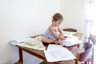 Young girl sitting on top of a wooden table covered in paperwork, holding pen, drawing. - MINF05411