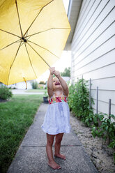Junges Mädchen im Sommerkleid steht auf einem Gartenweg neben einem Haus und hält einen gelben Regenschirm. - MINF05412