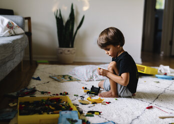 Junge mit braunen Haaren sitzt auf dem Boden in einem Kinderzimmer und spielt mit Lego. - MINF05418