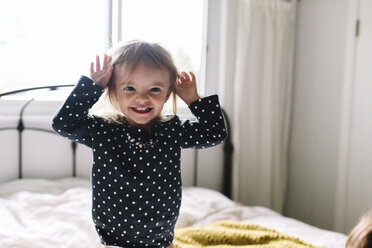 Junges Mädchen mit gepunktetem Pullover sitzt auf dem Bett, Hände auf dem Kopf, lächelnd. - MINF05424