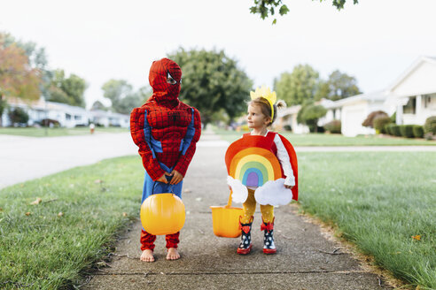 Junges Mädchen, das auf einem Bürgersteig steht und ein buntes Kostüm mit Regenbogen, Sonne und Wolken trägt, blickt auf einen Jungen im Spiderman-Kostüm, der neben ihr steht. - MINF05428