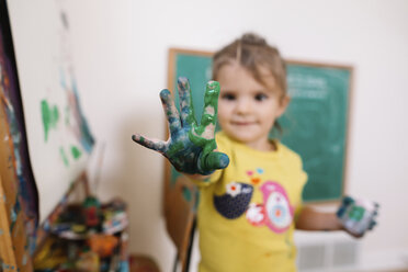 Ein junges Mädchen steht neben einer Staffelei und hält eine mit grüner und blauer Farbe beschmierte Hand in Richtung Kamera. - MINF05434