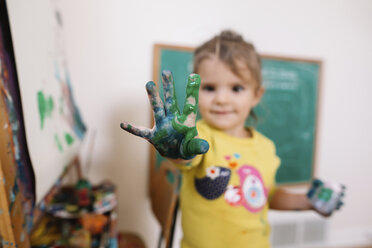 Young girl standing next to an easel, holding hand covered in green and blue paint up towards camera. - MINF05434