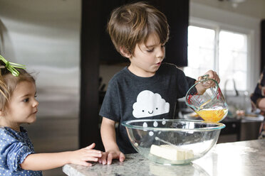 Junges Mädchen mit grüner Schleife im Haar steht in einer Küche und beobachtet einen Jungen, der Eier in eine Glasschüssel gießt. - MINF05443