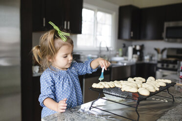 Junges Mädchen mit grüner Schleife im Haar steht in einer Küche, hält einen Löffel in der Hand und verziert Kekse. - MINF05444