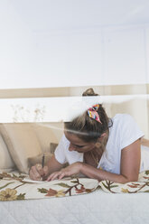 Young woman lying on bed at home writing in diary - KKAF01410