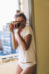 Porträt einer glücklichen jungen Frau, die am Fenster steht und mit einer altmodischen Kamera fotografiert - KKAF01424