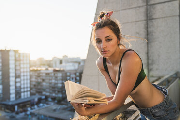 Portrait of young woman wearing bra reading book on balcony - KKAF01432