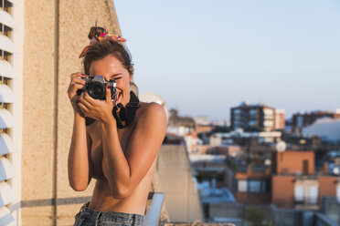 Happy topless young woman with camera taking pictures on balcony - KKAF01434