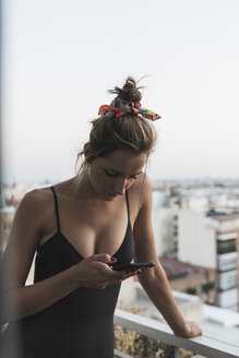 Junge Frau im schwarzen Kleid mit Handy auf dem Balkon - KKAF01453