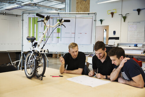 Three men in a meeting at a bicycle factory, sitting at a table with a folding bicycle on the table top. - MINF05473