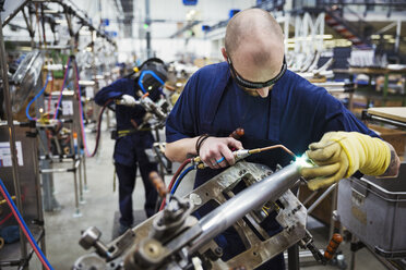 Männlicher Fabrikarbeiter mit Schutzbrille, der ein Schweißgerät benutzt und in einer Fabrik an einem Fahrrad arbeitet. - MINF05477