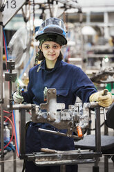 Weibliche Fabrikarbeiterin, eine junge Frau mit hochgezogenem Mundschutz, die ein Schweißgerät hält und an Fahrradrahmen arbeitet. - MINF05479