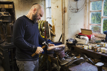 Ein Mann steht in einer Schuhmacherwerkstatt am Fenster und bearbeitet mit einem Hammer die Sohle eines Arbeitsstiefels. - MINF05492