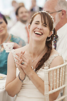 Die Braut in ihrem Hochzeitskleid sitzt in einem Festzelt, die Hände ineinander verschränkt und lacht. - MINF05506