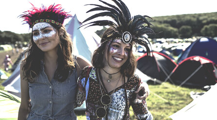Zwei lächelnde junge Frauen auf einem Sommermusikfestival mit geschminkten Gesichtern und Federkopfschmuck, die zwischen Zelten stehen. - MINF05552
