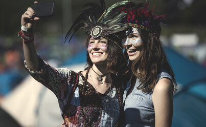 Zwei junge Frauen auf einem Sommer-Musikfestival mit bemalten Gesichtern und Federkopfschmuck, die ein Selfie mit ihrem Smartphone machen. - MINF05555