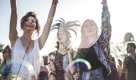 Drei lächelnde junge Frauen auf einem Sommer-Musikfestival mit geschminkten Gesichtern, Federkopfschmuck und erhobenen Armen, die in der Menge stehen. - MINF05557