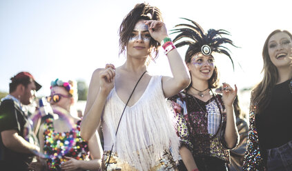 Two smiling young women at a summer music festival face painted, wearing feather headdress, standing among the crowd, looking at camera. - MINF05558