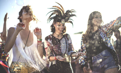Drei lächelnde junge Frauen auf einem Sommer-Musikfestival mit geschminkten Gesichtern und Federkopfschmuck, die in der Menge tanzen. - MINF05560