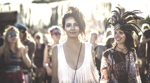 Two smiling young women at a summer music festival face painted, wearing feather headdress, standing among the crowd, looking at camera. - MINF05561