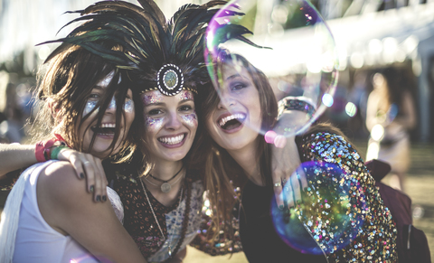 Three young women at a summer music festival wearing feather headdress and faces painted, smiling at camera. stock photo