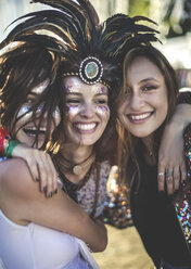 Three young women at a summer music festival wearing feather headdress and faces painted, smiling at camera. - MINF05566