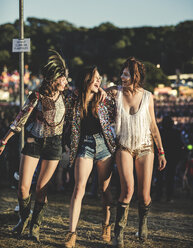 Drei junge Frauen auf einem Sommermusikfestival in Hotpants und Gummistiefeln, mit Federschmuck und bemalten Gesichtern, die Arme um die Schulter gelegt und lächelnd. - MINF05571