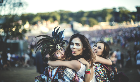 Drei junge Frauen auf einem Sommermusikfestival mit Federschmuck und bemalten Gesichtern, die in die Kamera lächeln und die Zunge herausstrecken., lizenzfreies Stockfoto