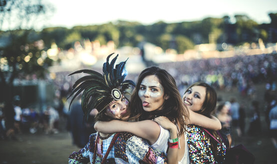 Drei junge Frauen auf einem Sommermusikfestival mit Federschmuck und bemalten Gesichtern, die in die Kamera lächeln und die Zunge herausstrecken. - MINF05572