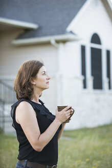 Eine Frau in einem ärmellosen Hemd, die eine Tasse in den Händen hält, steht vor einem Landhaus. - MINF05586