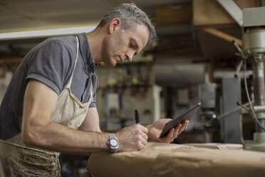 Ein Mann in einer Möbelrestaurierungswerkstatt benutzt ein digitales Tablet und schreibt auf ein in braunes Verpackungspapier eingewickeltes Paket. - MINF05589