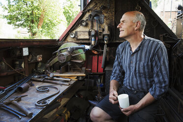 Blacksmith sitting on a working narrowboat on a waterway, in his workshop holding mug. - MINF05596