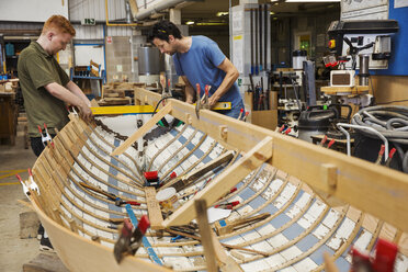 Zwei Männer in einer Bootsbauerwerkstatt, die gemeinsam an einem hölzernen Bootsrumpf arbeiten. - MINF05603