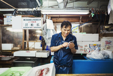 A traditional fresh fish market in Tokyo. A man in a blue apron standing behind the counter of his stall, using a smart phone covered with protective plastic. - MINF05690