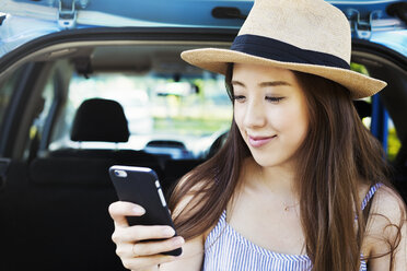 Lächelnde junge Frau mit langen braunen Haaren, die einen Panamahut trägt und ein Mobiltelefon benutzt. - MINF05755