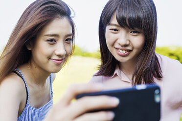 Zwei lächelnde junge Frauen mit langen braunen Haaren, die ein Mobiltelefon in der Hand halten und ein Selfie machen. - MINF05757