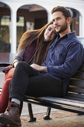 Ein junger Mann und eine junge Frau, die sich umarmen, sitzen auf einer Bank in einer städtischen Umgebung. - MINF05769