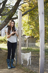 Eine junge Frau schaut auf ihr Handy neben einem Ziegenbock in einem Pferch. - MINF05787