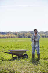 A young man in working clothes standing in a crop field with arms folded next to a wheelbarrow. - MINF05794
