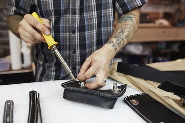 A leather worker and craftsman using a hand held tool on a blue leather bag on a workbench - MINF05801