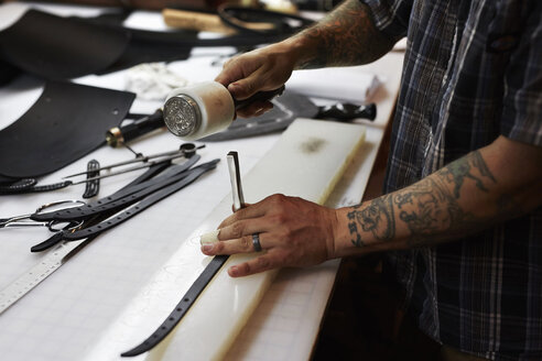 Ein Mann arbeitet an einer Werkbank in einer Lederwerkstatt und benutzt einen Hammer und einen Meißel, um Leder zu markieren. - MINF05802
