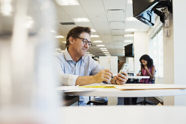 Ein Mann sitzt an einem Tisch in einem Büro und schaut auf ein Handy. - MINF05815