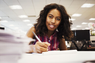 Eine Frau sitzt an einem Schreibtisch in einem Büro, hält einen Stift in der Hand und schaut in die Kamera. - MINF05819