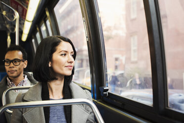 Eine junge Frau, die in einem Zug sitzt und aus dem Fenster auf eine Stadtlandschaft schaut, während ein Mann hinter ihr sitzt und wegschaut. - MINF05837