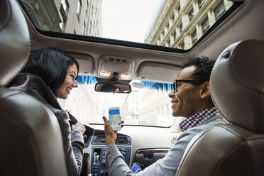 Eine junge Frau und ein junger Mann sitzen in einem Auto und betrachten eine Landkarte auf dem Display eines Mobiltelefons, gesehen vom Rücksitz aus. - MINF05839