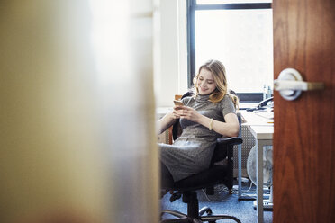 Eine Frau, die in einem Büro sitzt und ihr Telefon überprüft, gesehen durch eine offene Tür. - MINF05851