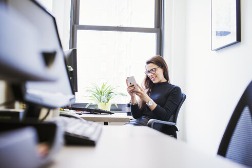 A young woman sitting at a desk in an office looking down at a cell phone. - MINF05864