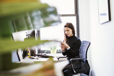 Eine junge Frau sitzt an einem Schreibtisch in einem Büro und schaut auf ein Handy. - MINF05866